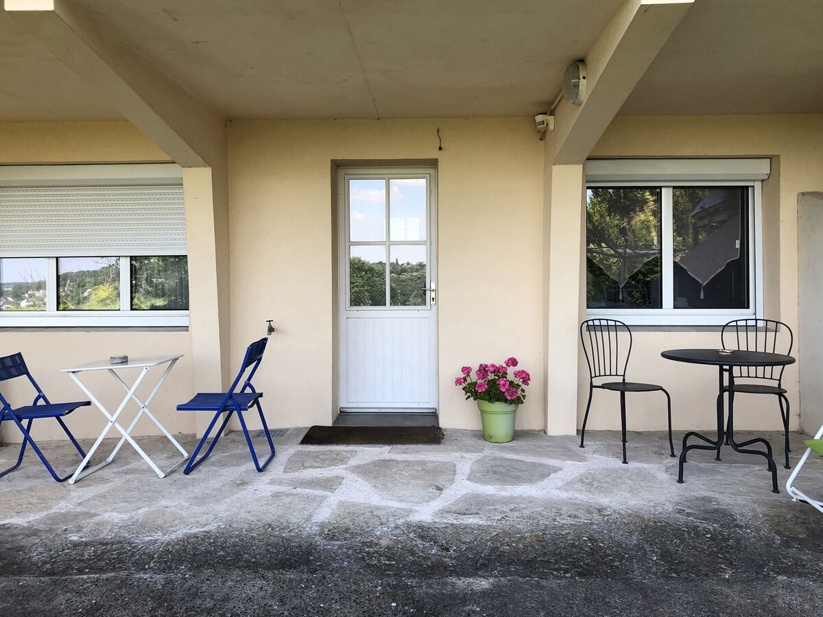 An outdoor area features a small table with two chairs alongside a second table and chair set. The entrance door is framed by two windows, with a potted plant adding a touch of greenery. The ground is made of stone, providing a clear passage.