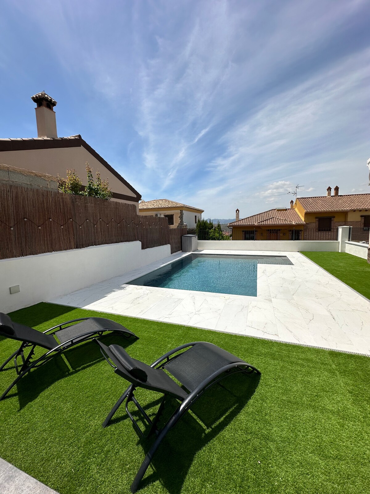 A private swimming pool is surrounded by a marble deck and lush green grass. Two black lounge chairs are positioned beside the pool, inviting relaxation. In the background, residential structures with tile roofs complement the serene outdoor setting under a bright blue sky.
