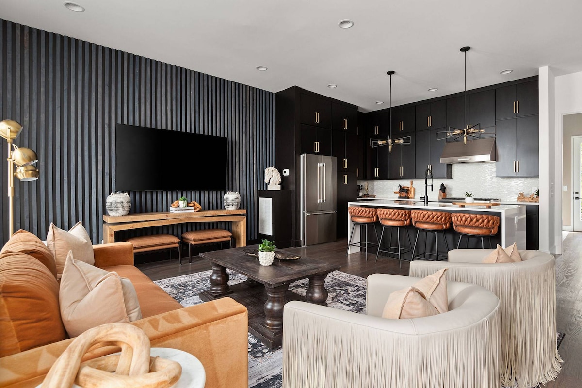 An open-concept living area showcases a stylish kitchen with modern appliances and seating at a spacious island. A comfortable seating arrangement features a mix of light and dark tones, with decorative accents and a large flat-screen TV mounted on a textured wall.