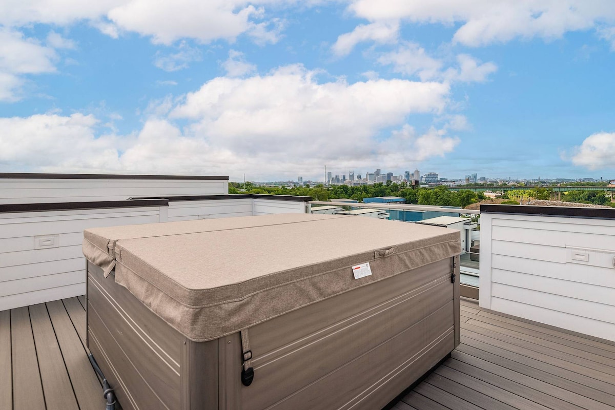 A hot tub is situated on a spacious rooftop deck, surrounded by wooden flooring and railings. In the background, city skyline views are presented against a bright, open sky.