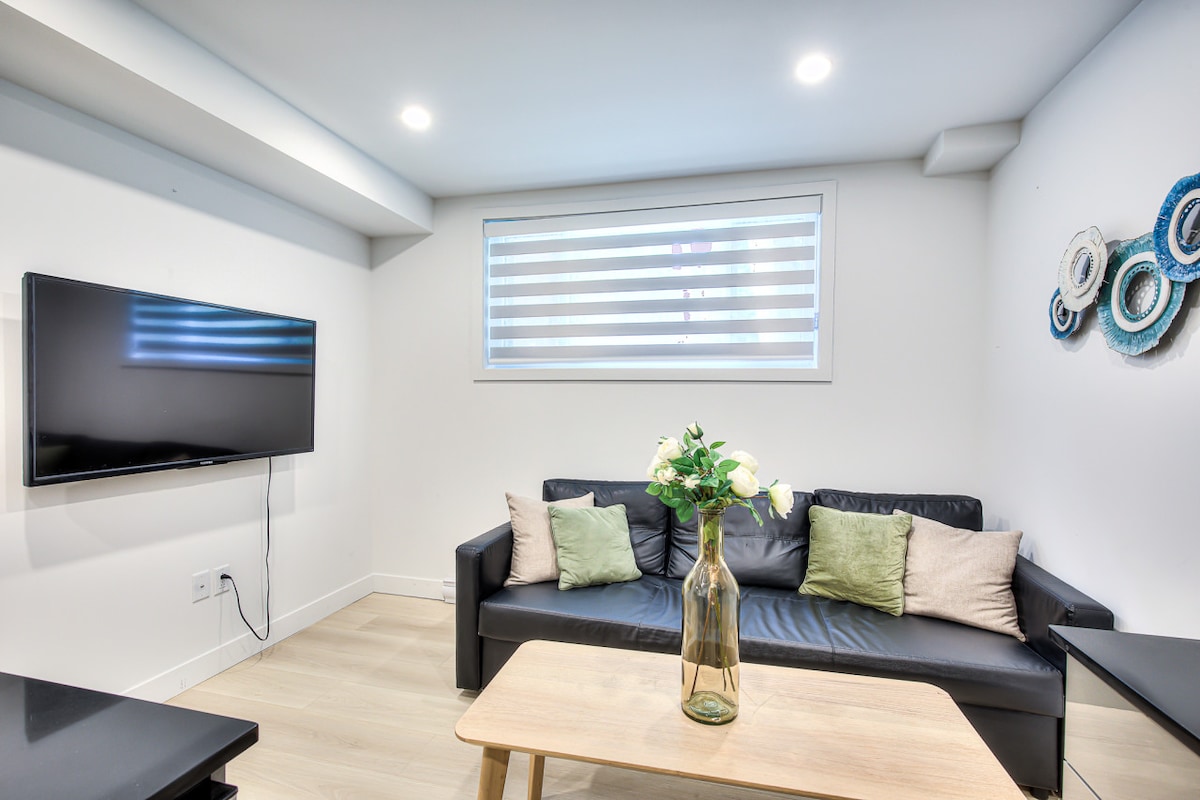A modern living room is presented, featuring a black sofa adorned with cushions in soft tones. A sleek coffee table sits in front, and a large flat-screen TV is mounted on the wall. Natural light filters through a window with striped shades.