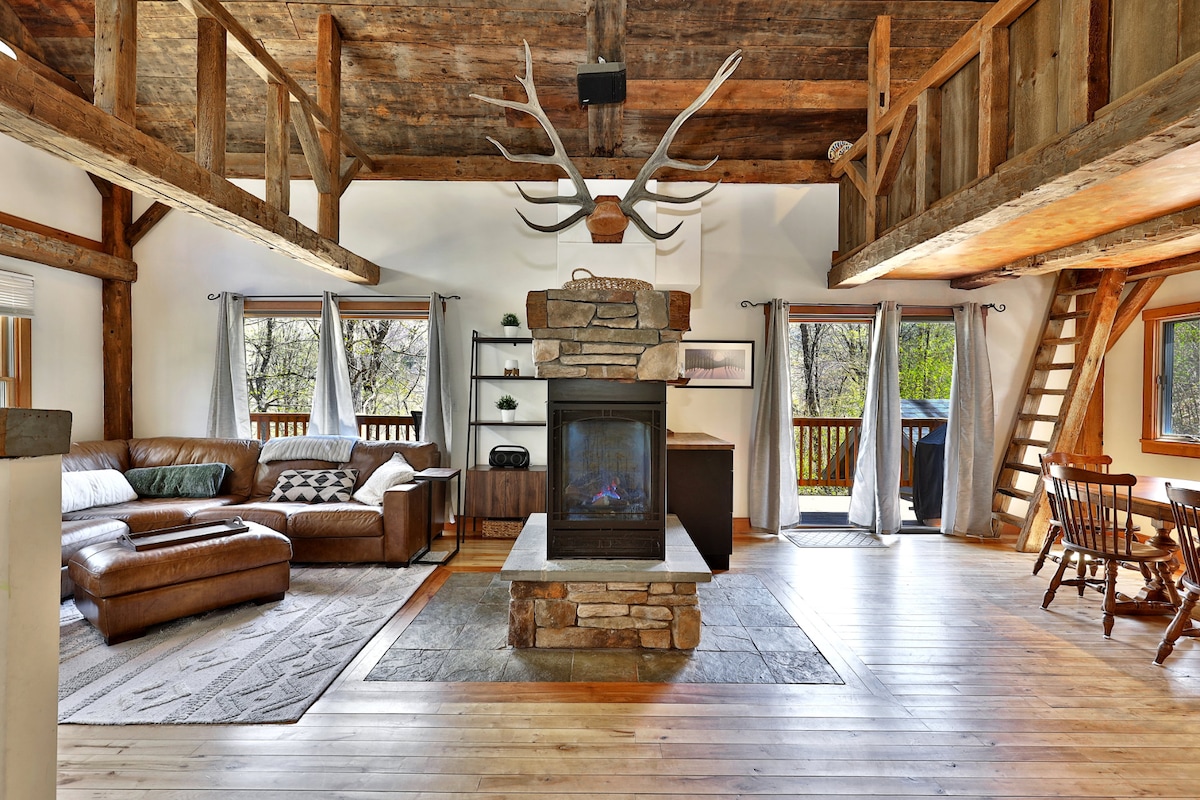 The spacious living area features a stone fireplace at its center, surrounded by a comfortable leather sectional and a rustic wooden dining set. Large windows provide natural light, and exposed wooden beams add to the cabin's character. A staircase leads to an upper loft area.