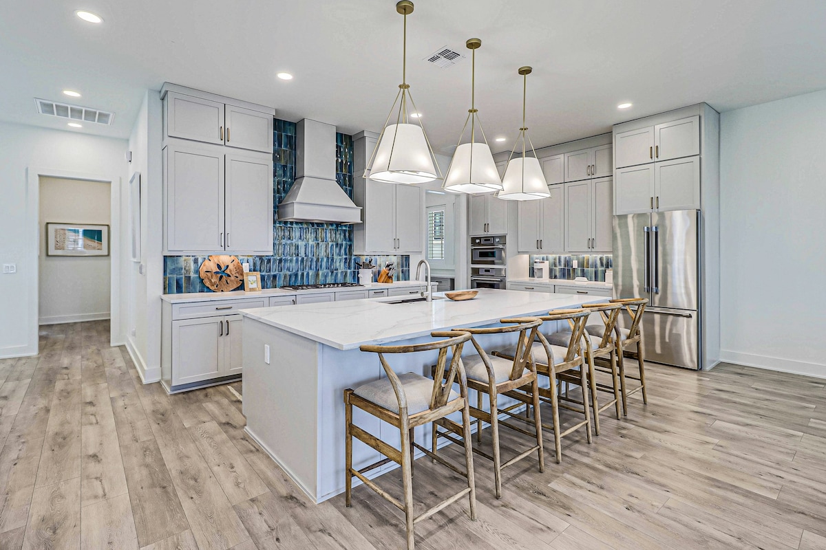 The kitchen features a spacious island surrounded by six light-colored chairs, enhancing the open layout. Sleek stainless steel appliances complement the modern cabinetry, while vibrant blue tile accents add a pop of color. Pendant lights illuminate the workspace, contributing to a bright and welcoming atmosphere.