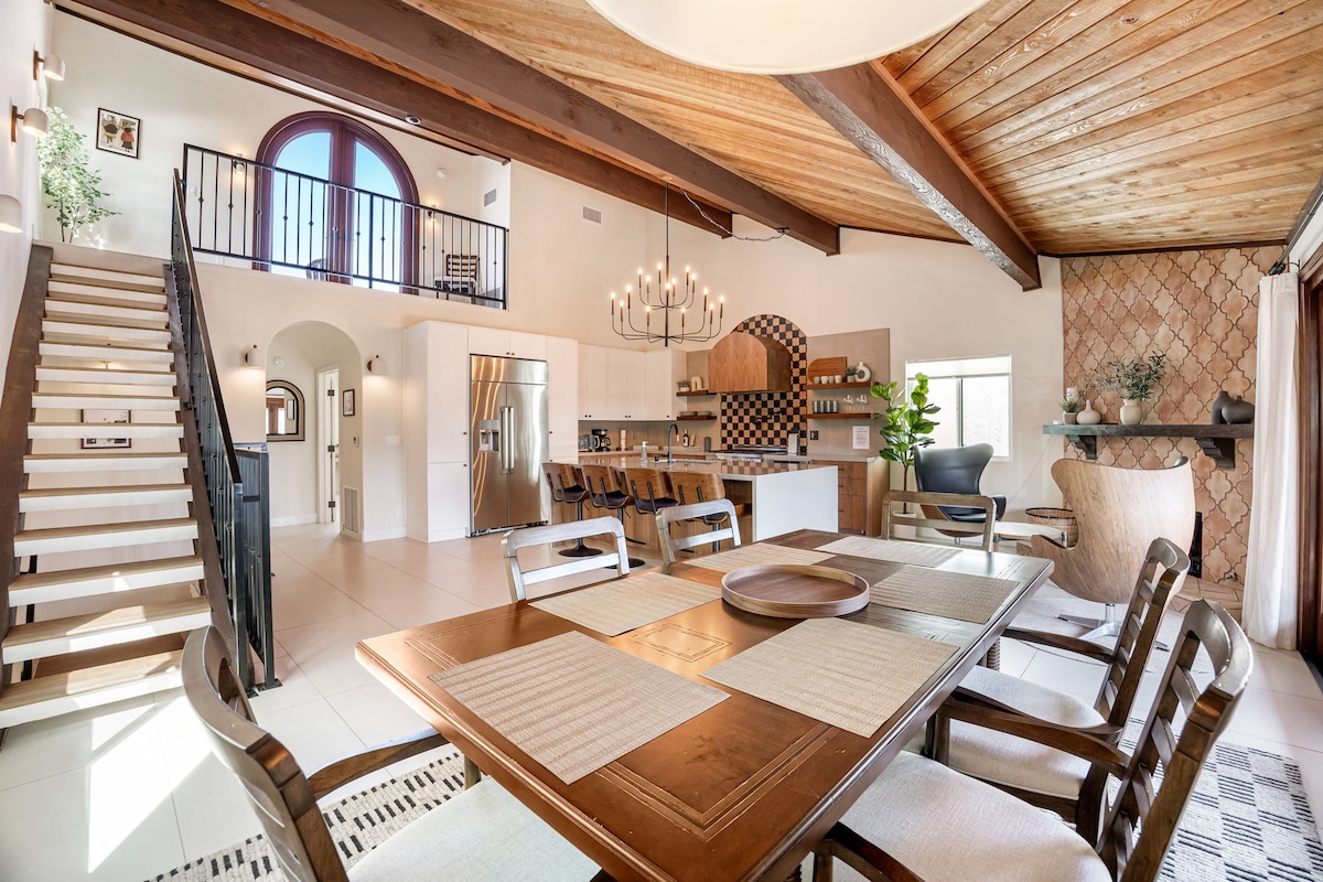 An open-concept dining area features a large wooden table surrounded by eight chairs. A modern chandelier hangs above, illuminating the space. In the background, stylish kitchen appliances and cabinetry are visible, along with a staircase leading to the upper level. Natural light fills the room through large windows.