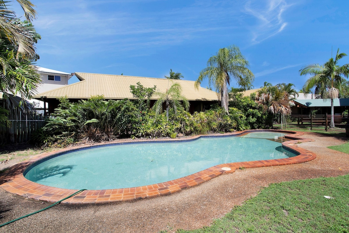 A spacious outdoor pool is surrounded by tropical greenery and features a curved shape with a textured brick border. A clear blue sky is visible above, enhancing the pool area’s inviting atmosphere.