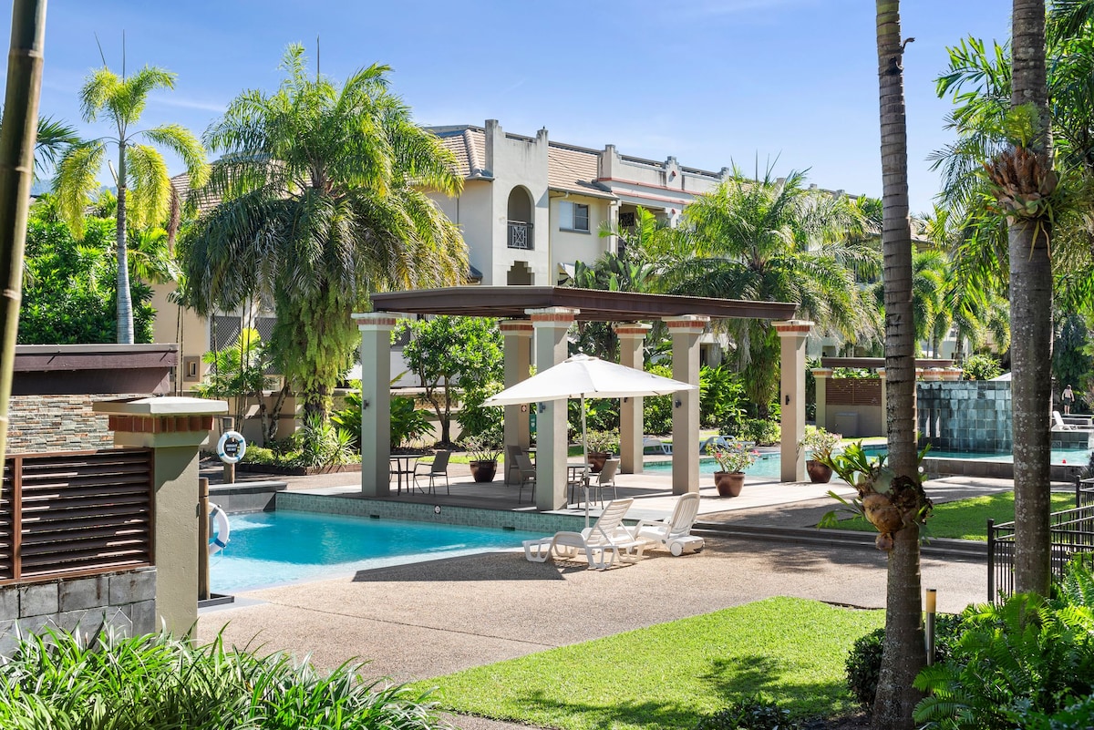 A serene pool area is surrounded by lush tropical landscaping, featuring a shaded seating area with a white umbrella. Sun loungers are positioned for relaxation, and a glimpse of additional pools and lush greenery is visible, creating a tranquil atmosphere.