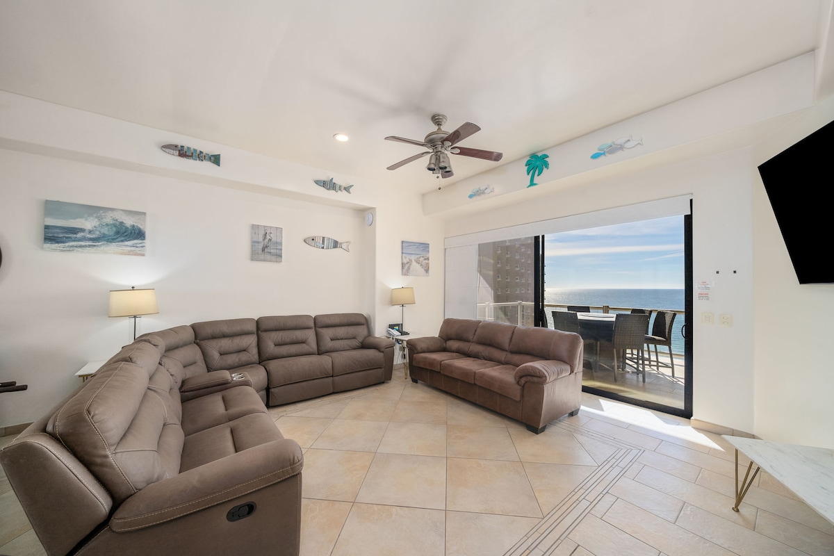 A spacious living area features comfortable seating arranged in a circular layout, with two large sofas and a recliner. Light-colored walls and tiled flooring enhance the room's brightness. Large glass doors open to a balcony offering ocean views, complemented by wall-mounted artwork.