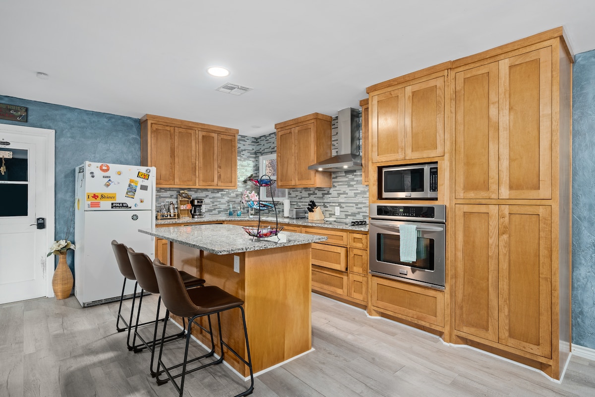 The kitchen features wooden cabinetry and a spacious granite island with seating for three. Modern appliances, including a stove and double oven, are present. A refrigerator adorned with magnets adds a personal touch. The blue walls create a calming contrast with the light-colored flooring.