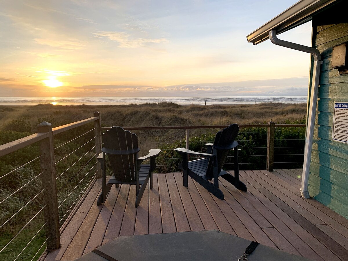 A spacious deck features two black Adirondack chairs, positioned to overlook the beach and ocean. The view captures a serene sunset with soft orange and pink hues reflecting on the water, complemented by grassy dunes in the foreground.