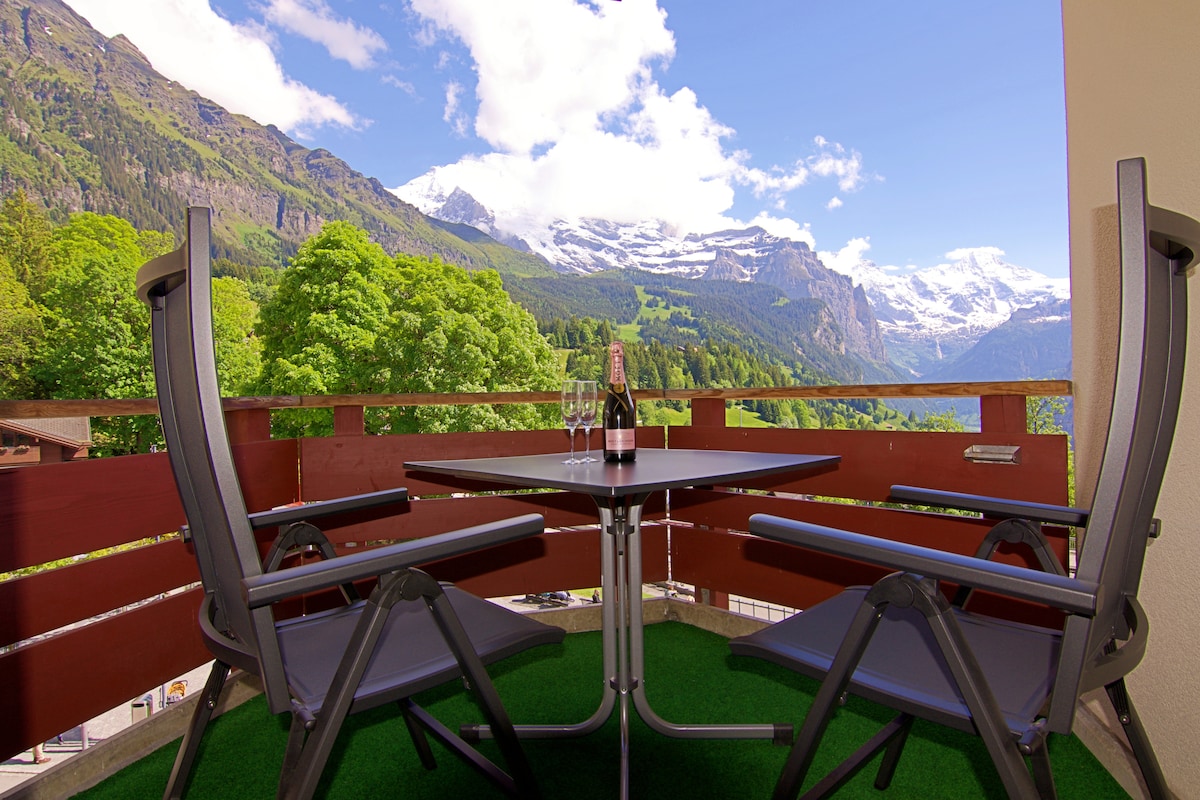 A private balcony features a round table with two chairs, offering a serene space for relaxation. Views of the surrounding mountains and lush greenery are visible, framed by the balcony's wooden railing. The floor is covered with a green outdoor rug, enhancing the comfortable atmosphere.