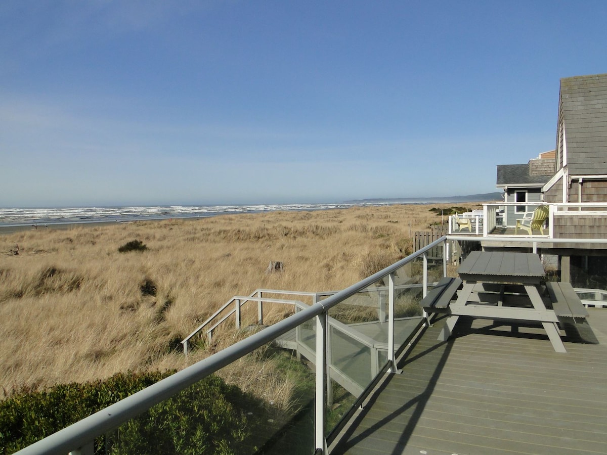 A spacious deck features a picnic table, offering unobstructed views of the ocean and surrounding dune grasses. Expansive beach stretches into the distance under a clear blue sky, creating a serene coastal atmosphere.