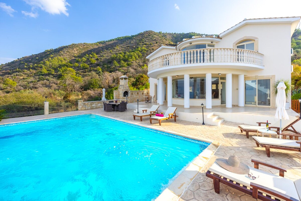 The villa exterior is showcased with a private pool and a sun terrace featuring loungers. A barbecue area is visible nearby. The backdrop features a gently sloping hill adorned with greenery, while large glass doors provide views of the pool and surroundings.