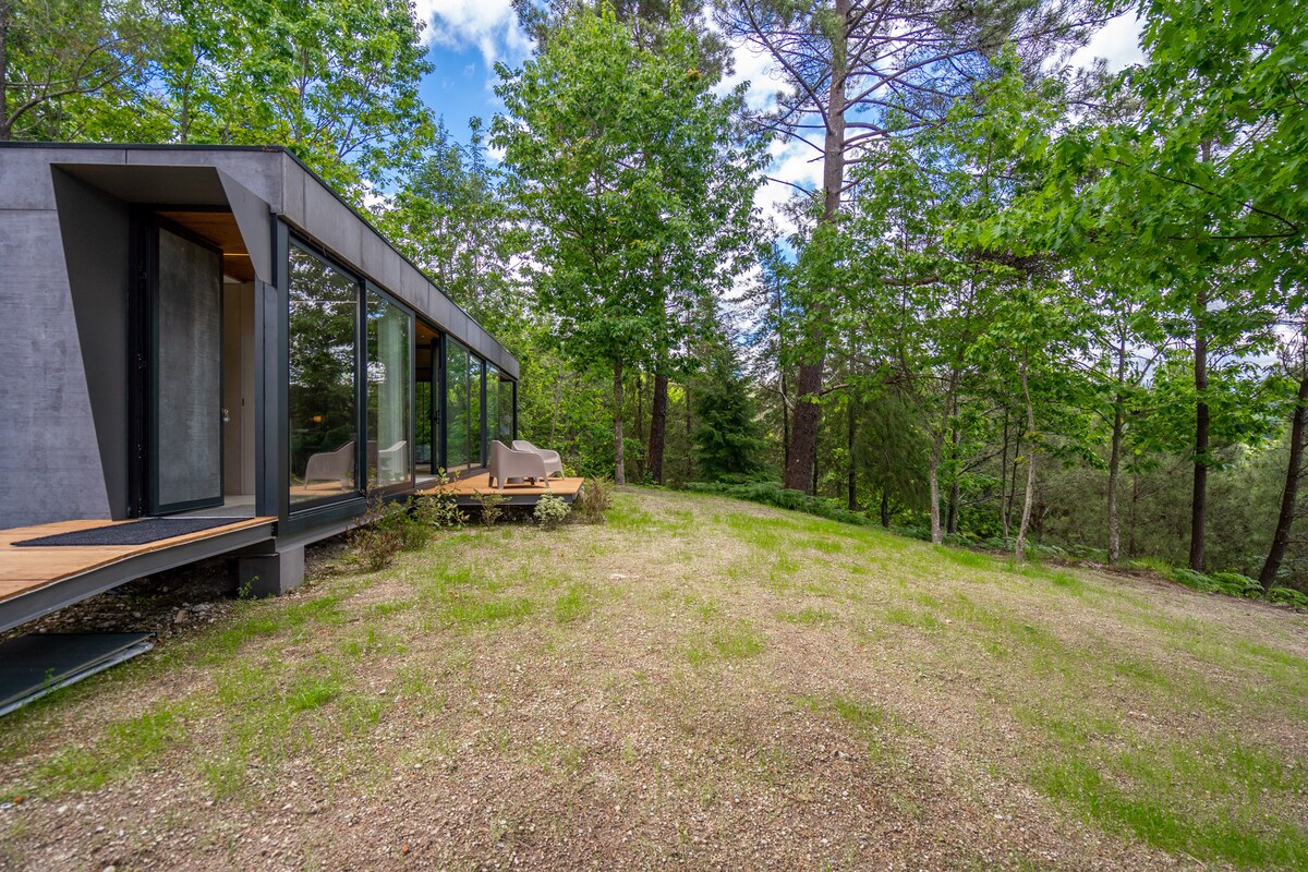 A modern villa is nestled among lush green trees, featuring large glass windows that create a seamless connection with nature. An outdoor seating area is visible on the wooden deck, offering a tranquil spot for relaxation amidst the natural surroundings.