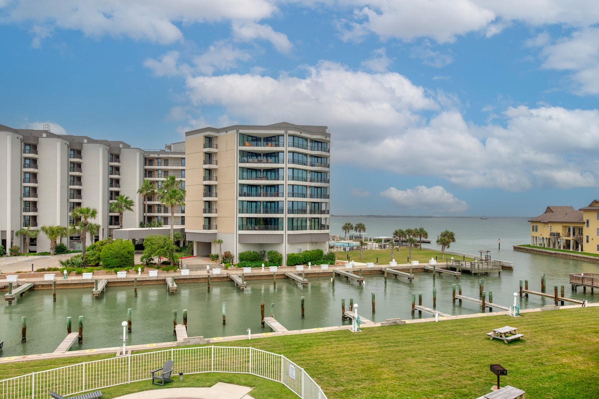 A view of the condo building from the waterfront, showcasing multiple boat slips along the canal. Lush greenery surrounds the marina, with an expansive lawn in the foreground and the coastal waters in the background, under a partly cloudy sky.