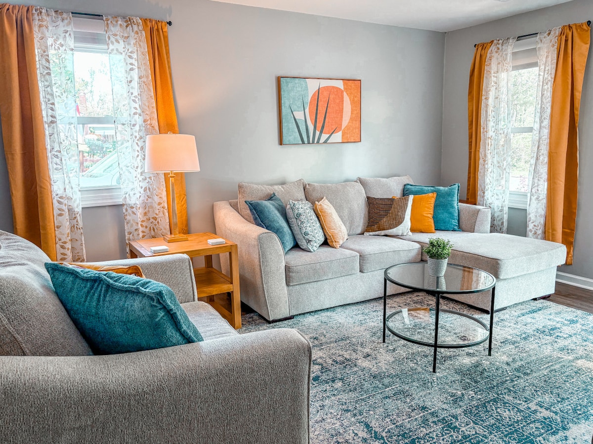A comfortable living area is presented with a gray sectional and matching chairs, enhanced by bright throw pillows. Natural light filters through the windows adorned with sheer curtains and orange drapes. A round coffee table sits centrally, accompanied by a decorative plant.