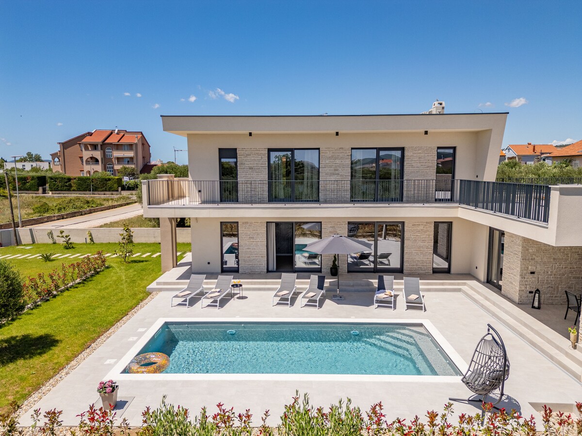 The villa's exterior showcases a two-story design with expansive glass doors and a spacious terrace overlooking a heated swimming pool. Comfortable loungers are placed around the pool, complemented by an umbrella for shade. A manicured lawn and flower beds frame the space, enhancing the vibrant outdoor setting.