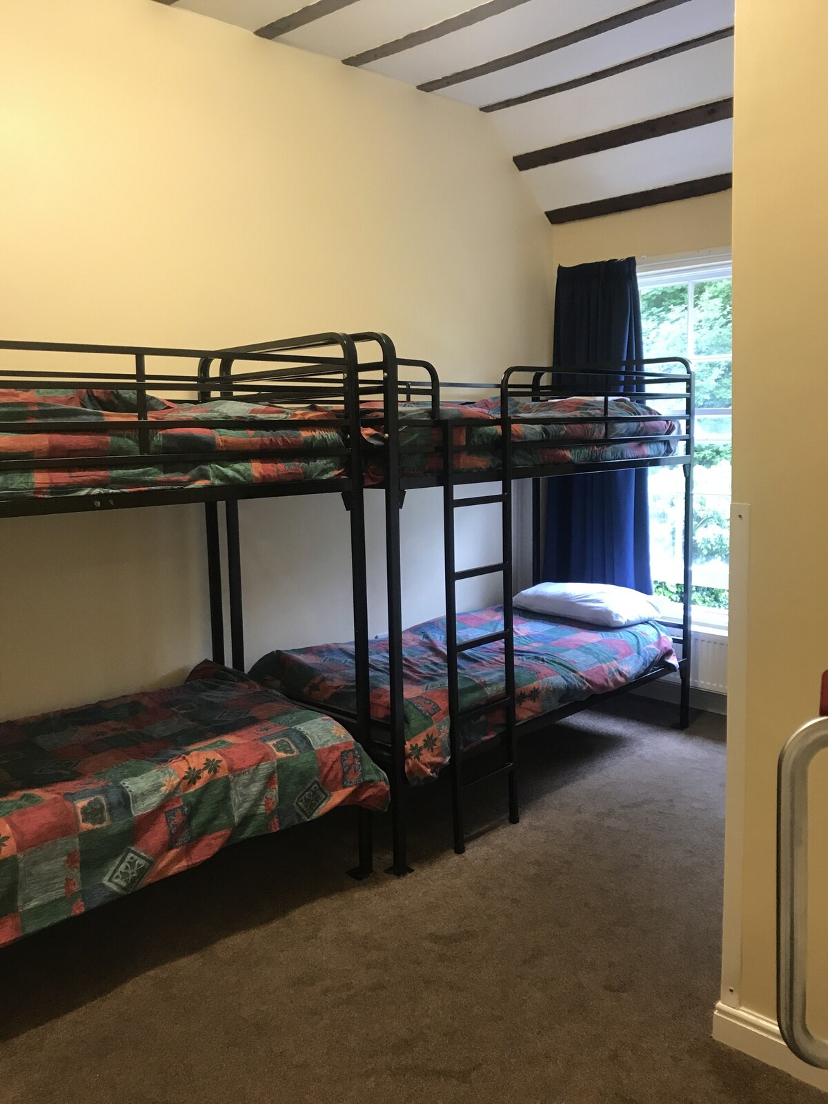 A room features two sets of sturdy metal bunk beds, each with colorful bedding. Natural light enters from a window, which is dressed with dark curtains. A carpeted floor provides comfort, while the room is minimally decorated for a clean and functional appearance.
