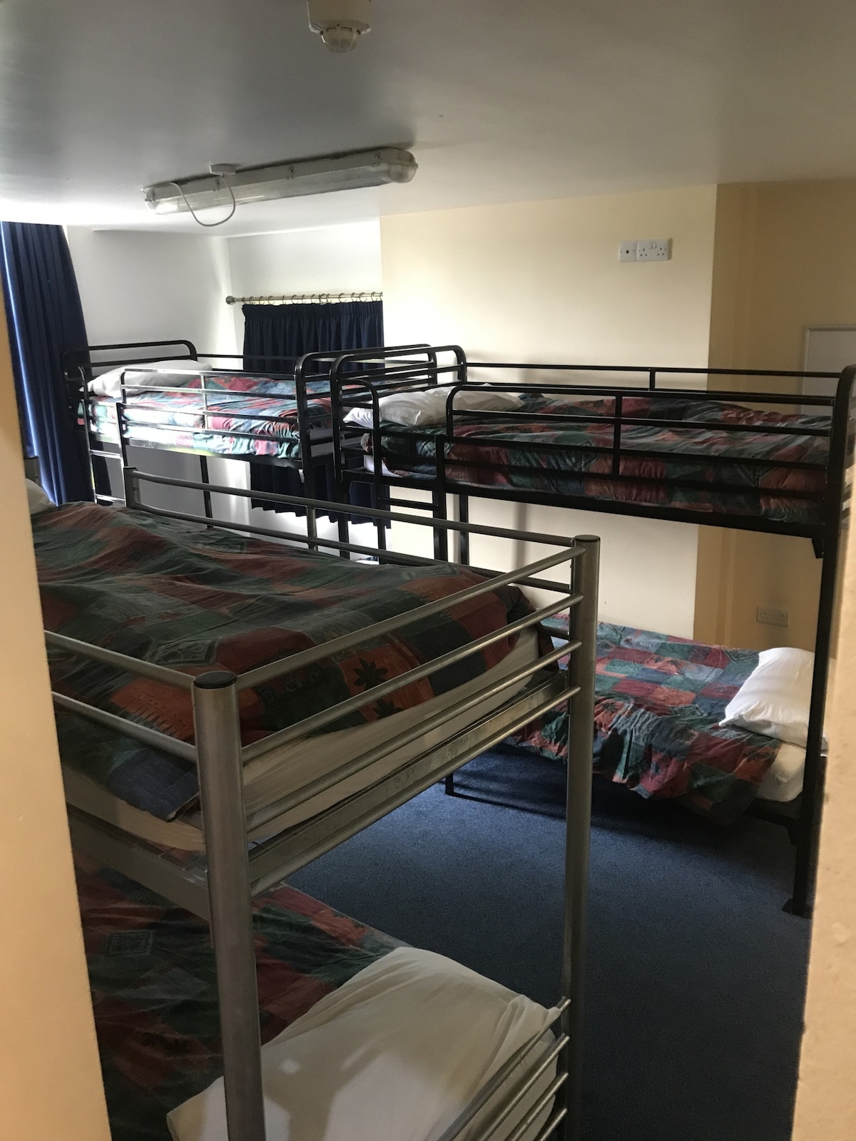 A room featuring four sets of bunk beds arranged in an efficiently designed layout. Colorful bed linens add a touch of vibrancy. Natural light filters through a window dressed with dark curtains, illuminating the space and highlighting the carpeted flooring.