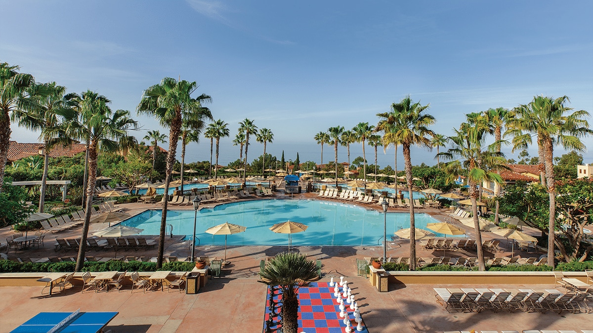 A large pool area is surrounded by palm trees and lounge chairs. Umbrellas provide shade over some seating areas. A vibrant blue sky complements the inviting swimming pool, while a colorful checkerboard area adds a playful touch to the scene.