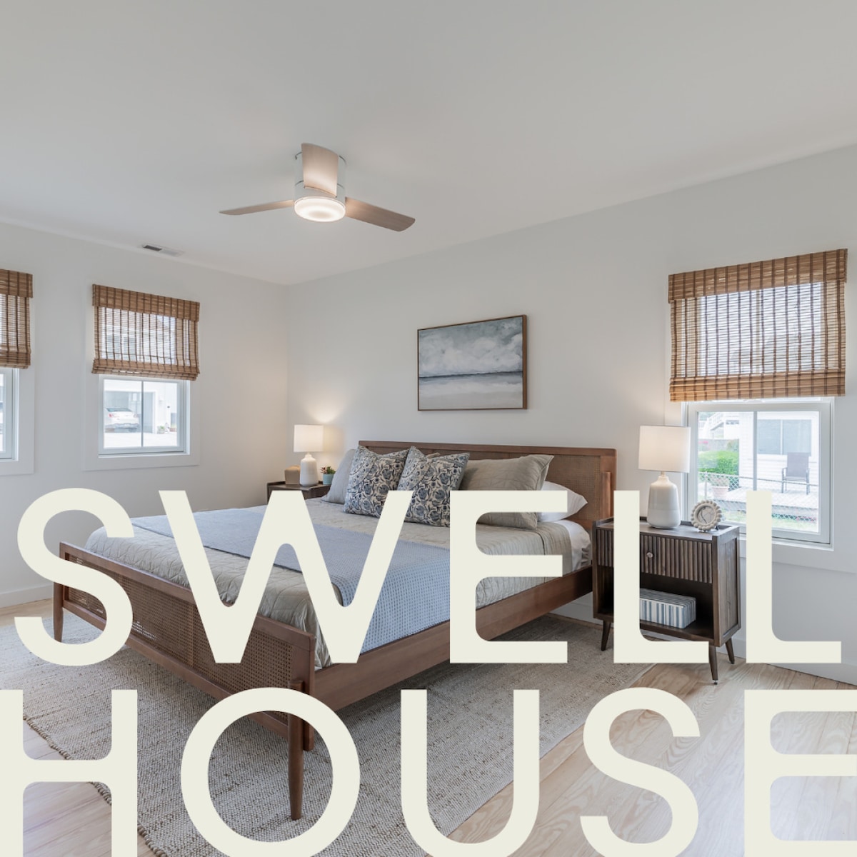 The primary bedroom features a spacious bed adorned with neutral bedding. Two bedside lamps illuminate the room, complemented by natural light from the tinted front windows. Light brown window shades provide privacy, and a piece of wall art adds a touch of character.