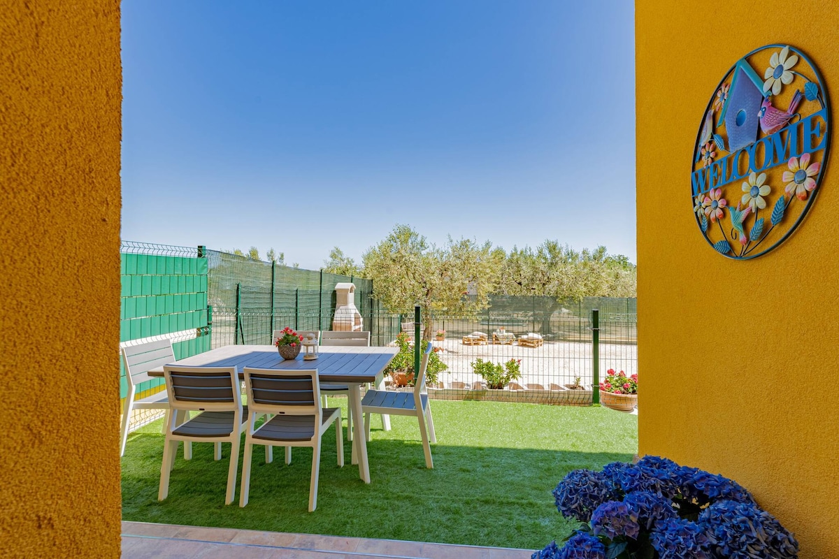 A welcoming outdoor area features a dining table with several chairs, set on green grass. Colorful flowers decorate the table, while a ‘Welcome’ sign adds a friendly touch. The fenced garden includes olive trees, contributing to a serene outdoor atmosphere.