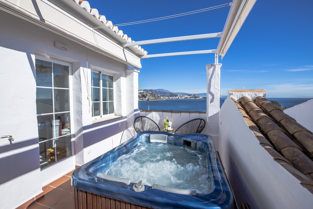 A private jacuzzi is positioned on the upper terrace, offering scenic views of the sea and mountains. Clear blue skies are visible above, while the surrounding area features white walls and a tiled roof, enhancing the tranquil setting.