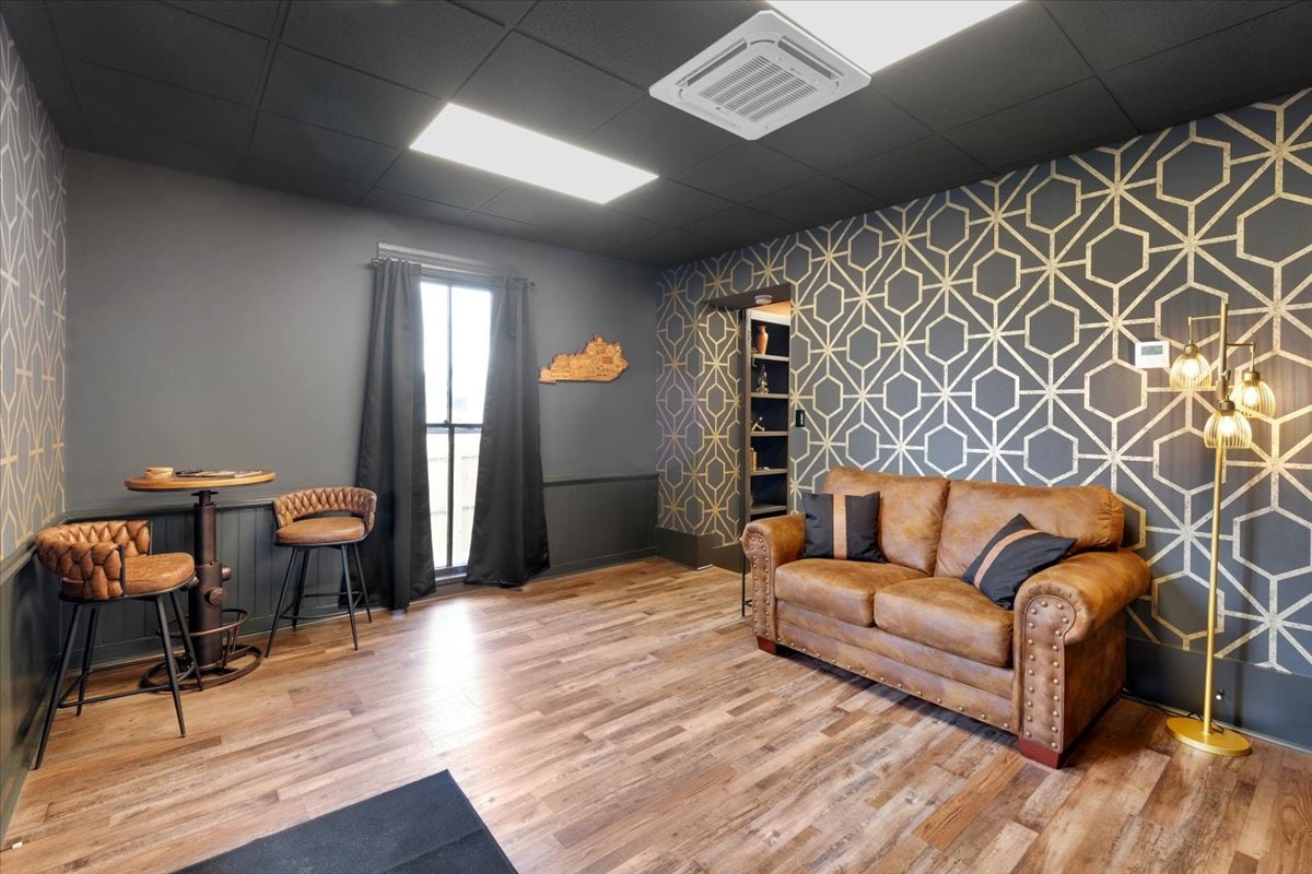 The living area features stylish geometric wallpaper against a dark backdrop. A brown leather sofa is complemented by two high-top chairs and a small table. Natural light filters through a window dressed in black curtains, illuminating the warm wood flooring.