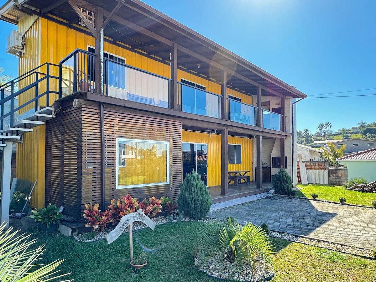 The exterior showcases a vibrant yellow two-story container structure, featuring large glass doors and a welcoming balcony. A well-maintained garden with decorative plants and a paved pathway create an inviting entrance, complemented by a sitting area and an outdoor dining table.