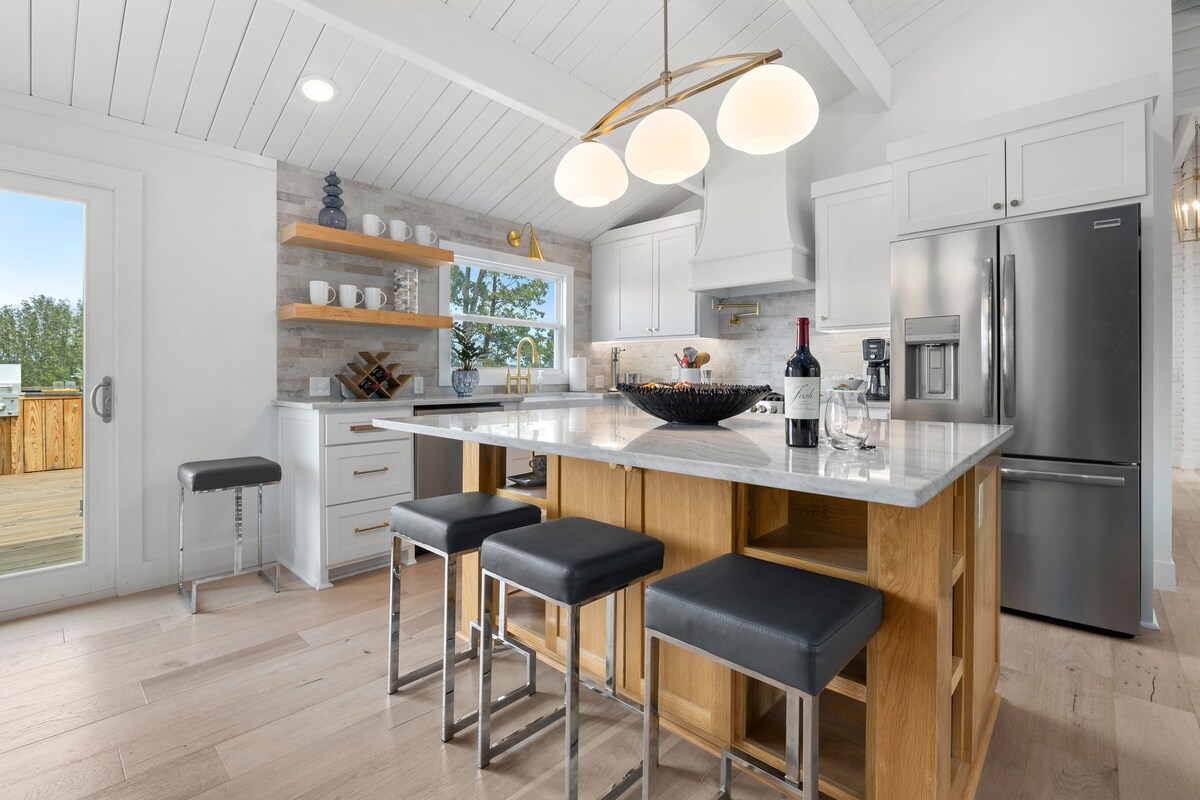 A modern kitchen features a large island with seating for three, complemented by sleek black stools. White cabinetry and open shelving showcase dishware and wine. Stainless steel appliances include a refrigerator and oven. Natural light floods the space through a door leading to the outdoor deck.