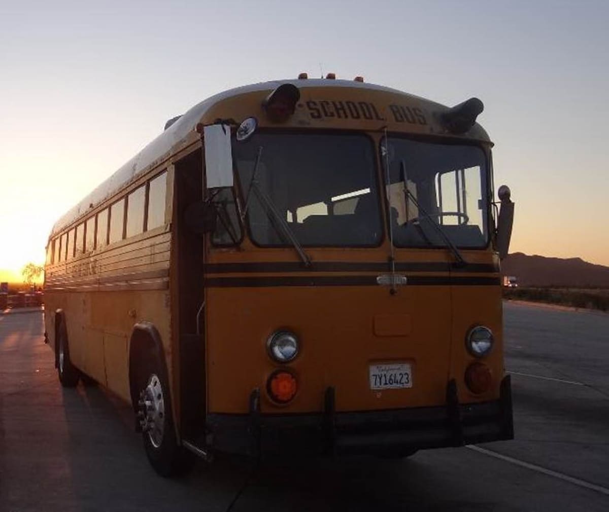 A classic yellow school bus is parked on a solid surface under a soft sunset glow. The bus features large windows, an elevated roofline, and distinctive black accents, emphasizing its nostalgic design and character. Mountains are visible in the background.