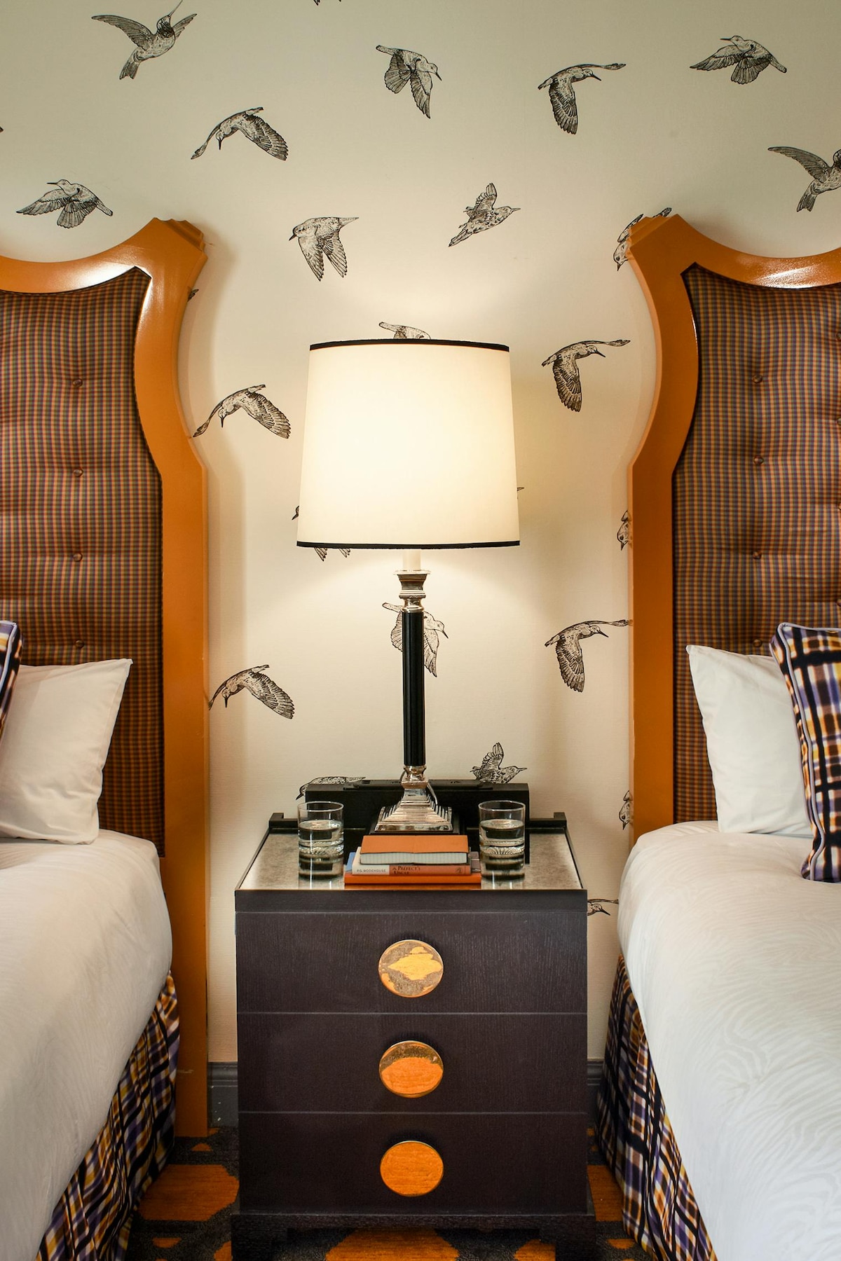 A well-appointed guest room features two spacious beds with tufted headboards flanking a modern bedside table. A stylish lamp casts light over the table, which is adorned with glasses. The wall displays a decorative pattern of birds for added character.