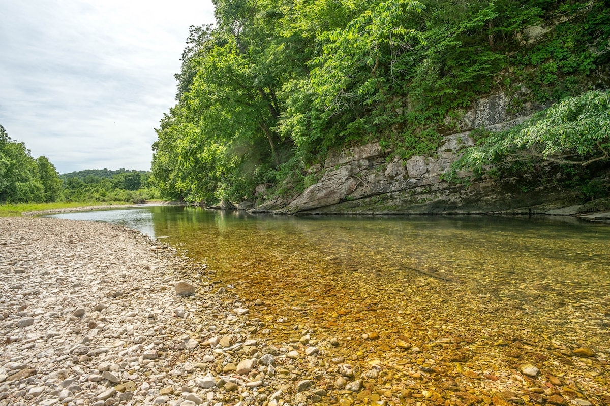 A clear creek flows gently past smooth stones and pebbles along the shoreline. Lush greenery and trees provide a natural backdrop, while the calm water reflects earthy tones and a muted sky.