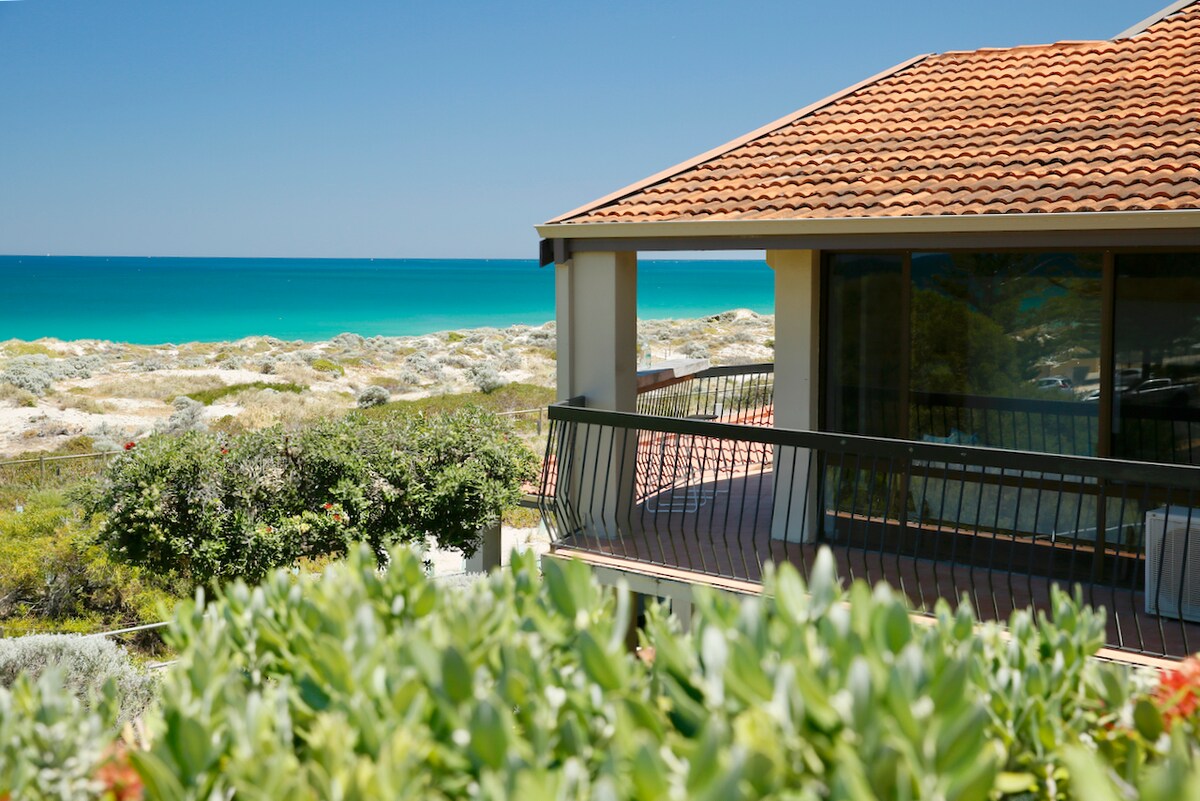 A coastal apartment is shown with a terracotta-tiled roof and expansive balcony overlooking the sparkling turquoise waters of the ocean. Lush greenery surrounds the property, outlining the views of the sandy beach beyond, creating a serene and inviting atmosphere.