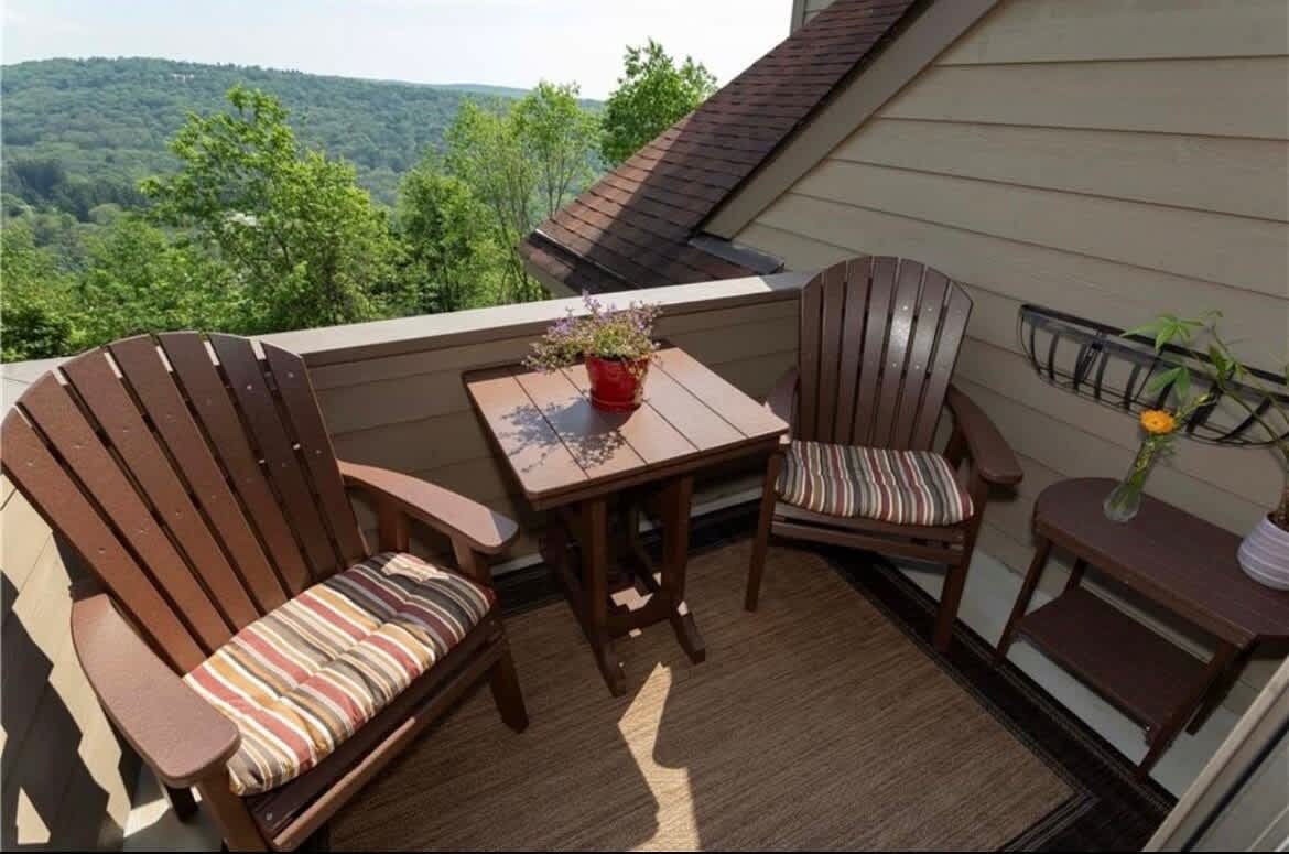 A small outdoor balcony features two wooden Adirondack chairs with striped cushions positioned around a square table. A potted plant and a small vase with flowers are placed on the table. Lush greenery and rolling hills are visible in the background.