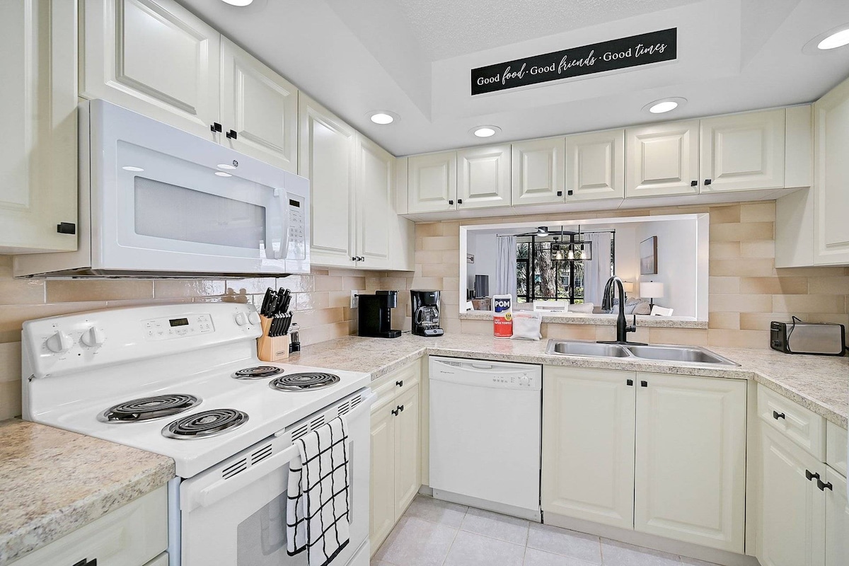 A bright and airy kitchen features cream-colored cabinetry and light countertops. Basic appliances, including a stove, microwave, dishwasher, and coffee maker, are neatly arranged. Under-cabinet lighting illuminates the workspace, while an open view leads to the living area, enhancing the connected layout.