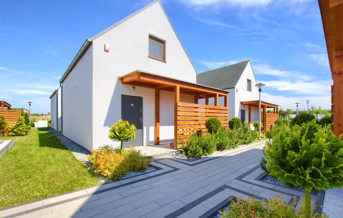The two-story holiday home features a modern exterior with light-colored walls and wooden accents. A landscaped garden with greenery surrounds the entrance, which is accessed via a stone pathway. Blue skies and gentle clouds provide a bright backdrop to the property.