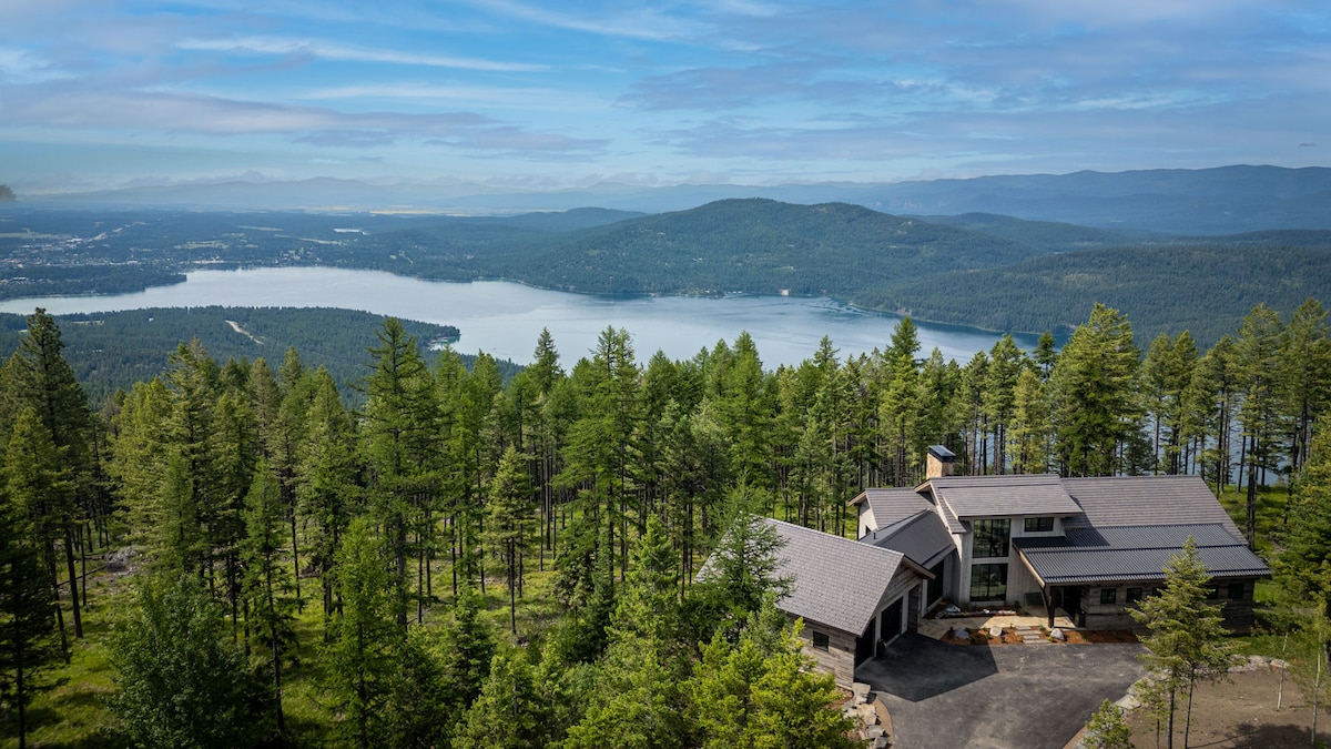 Aerial view of a modern mountain home nestled among lush, green trees, overlooking a large lake and mountains in the distance under a blue sky with scattered clouds. The exterior is designed with a combination of stone and wood, blending seamlessly into the natural surroundings.