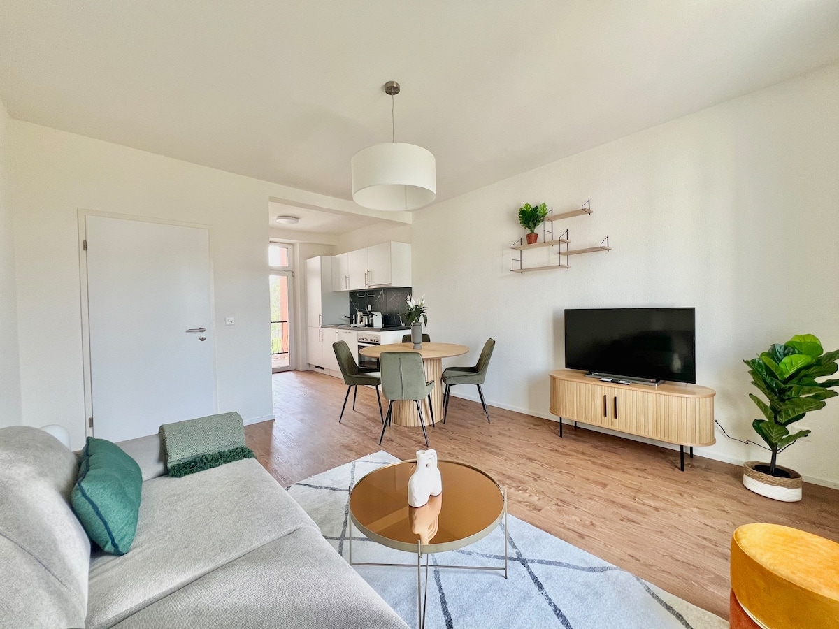 A contemporary living area is observed featuring a grey sofa and a round coffee table. A dining space with four green chairs is visible, along with a flat-screen television mounted on a wooden console. A potted plant adds a touch of greenery to the setting.