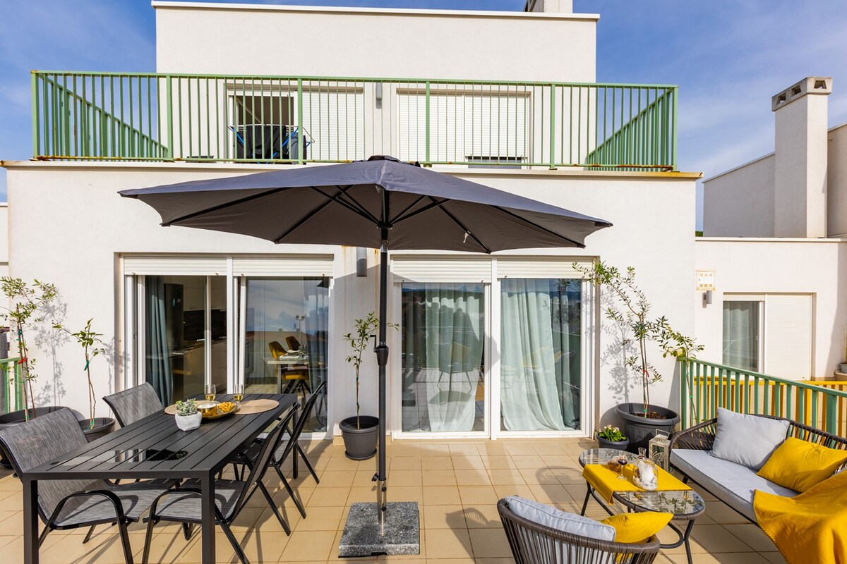 A large sunbathed balcony is featured, furnished with a dining table and chairs under a large umbrella. Cozy seating is positioned to the side, surrounded by decorative plants, providing a spacious area for relaxation and outdoor dining.