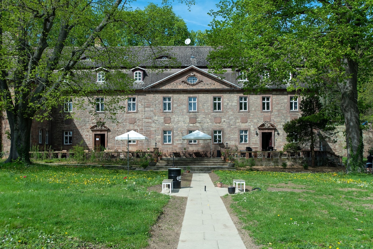 The historical building features a stone façade with several windows and a central entrance. Two outdoor seating areas are provided under white umbrellas, surrounded by a grassy area dotted with wildflowers. A walkway leads from the foreground to the entrance, framed by large trees.
