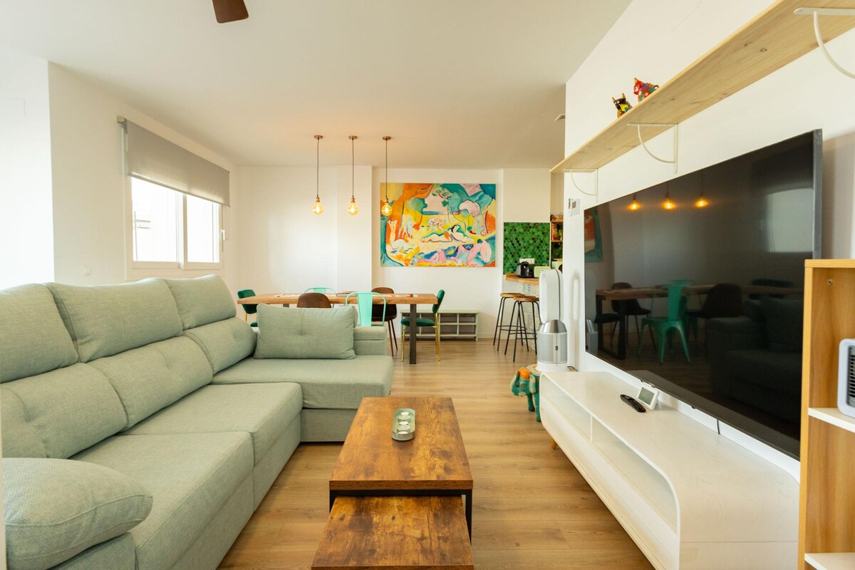 A spacious living area features a comfortable light green sofa and a wooden coffee table, complemented by a large flat-screen television mounted on the wall. Bright overhead pendant lights illuminate the space, while a colorful artwork adds a cheerful touch to the room.