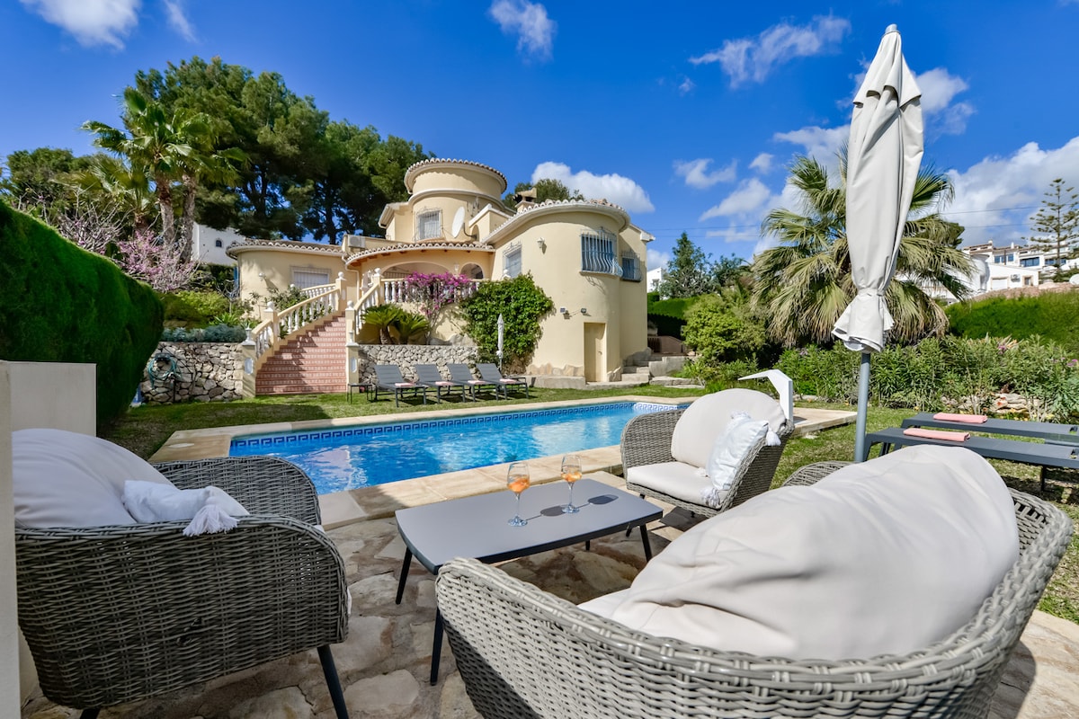 A seating area featuring two comfortable chairs and a table is positioned near a pool. The pool area is surrounded by lush greenery and palm trees, offering a serene ambiance. A villa stands in the background, complemented by clear blue skies and scattered clouds.