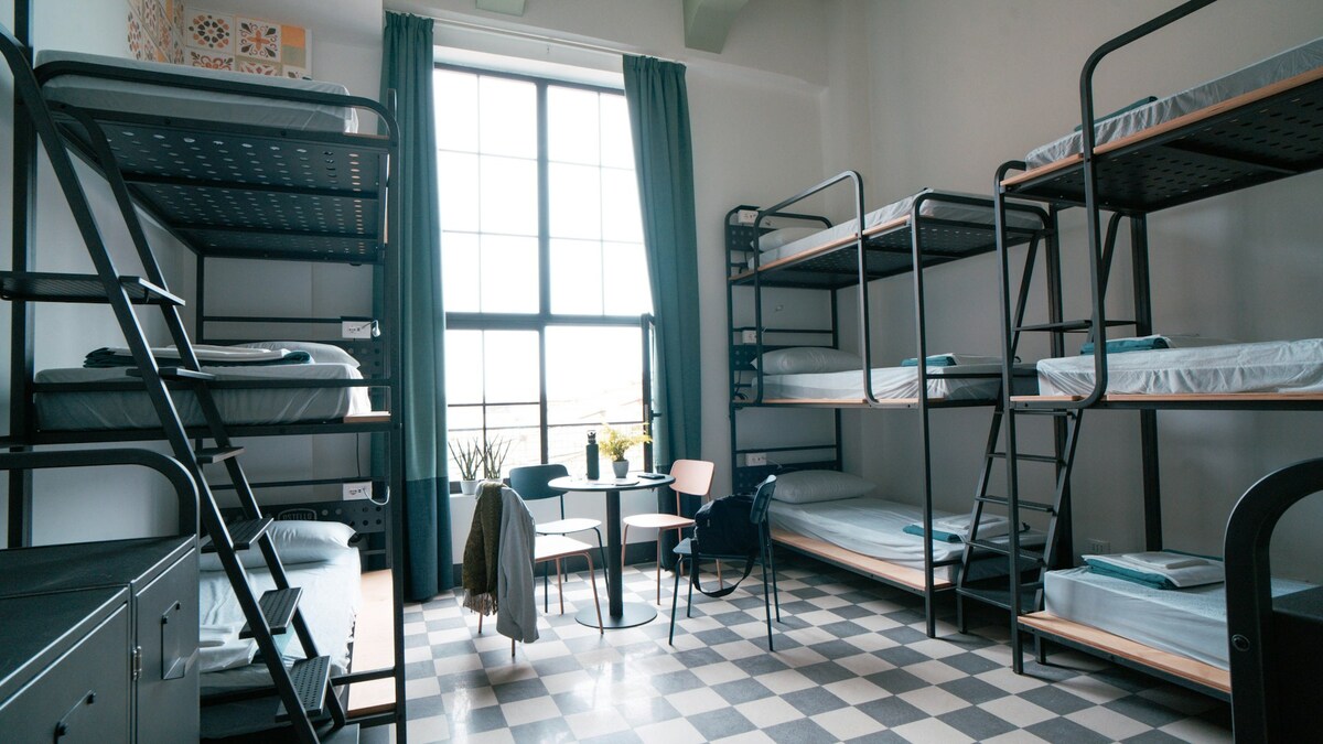 A spacious room features multiple bunk beds arranged along the walls, with bright windows allowing natural light to fill the space. A small round table with chairs sits centrally, and dark green curtains frame the windows, complementing the checker-patterned floor.