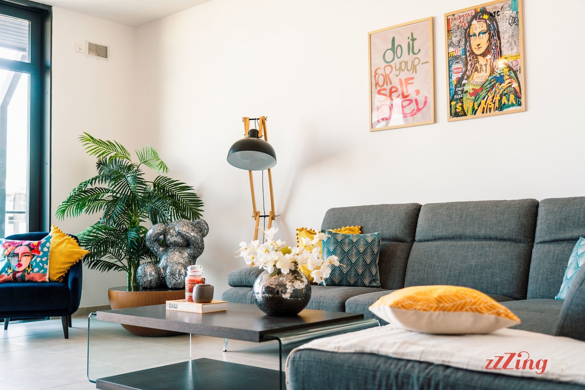A stylish living room features a modern gray sofa adorned with colorful cushions. A coffee table is centered on the light-colored floor, topped with decorative items, including a vase of flowers. A vibrant green plant adds freshness to the space, alongside framed art on the walls.