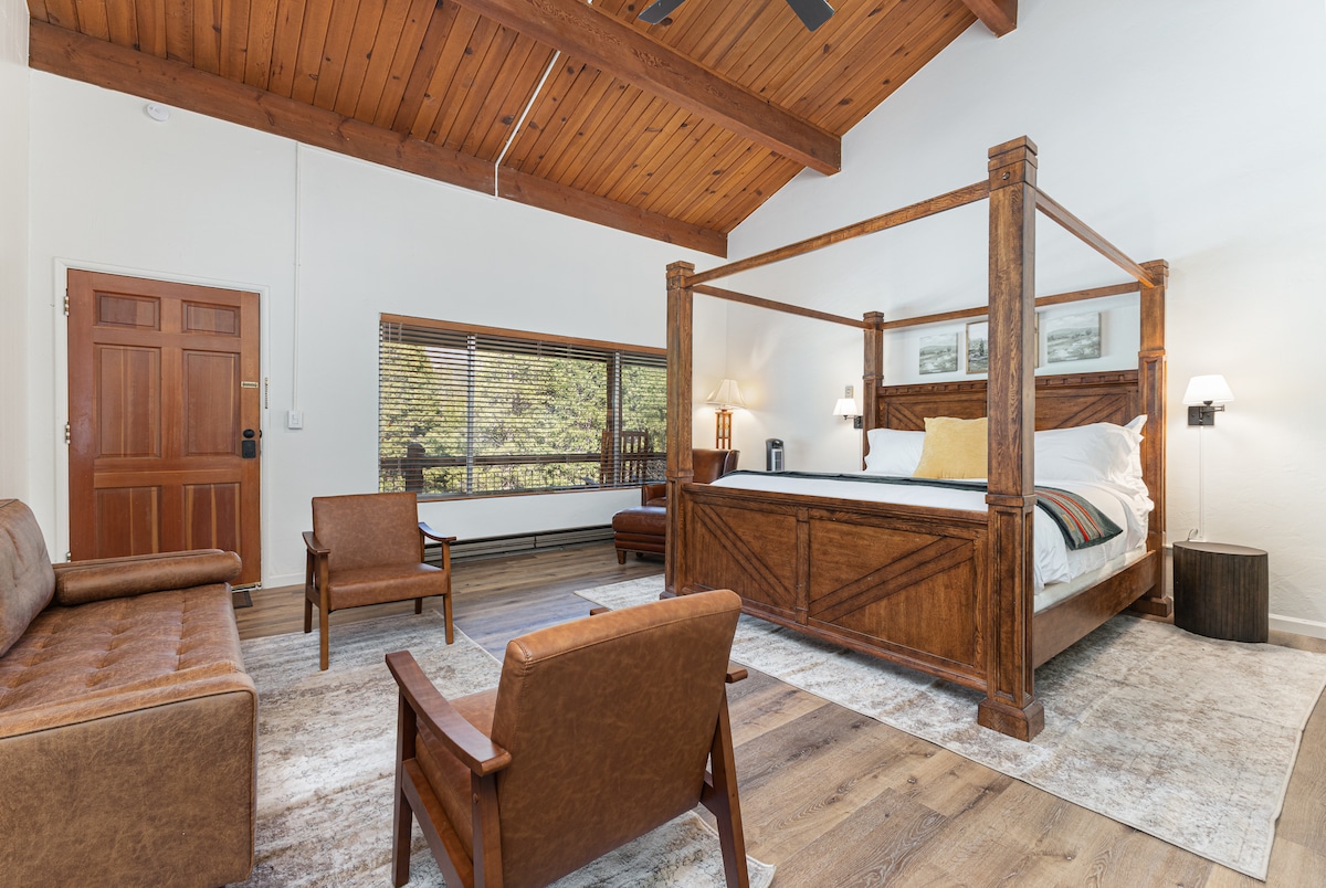 A spacious room features a king canopy bed crafted with wooden beams, complemented by modern furniture including two brown leather chairs and a brown sofa. Natural light filters through the window, illuminating the serene space with wood-paneled ceiling and warm wooden accents.