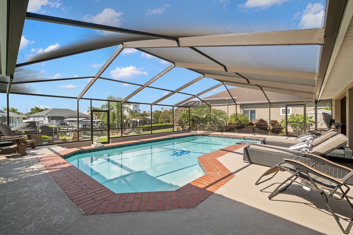 A screened-in pool area features a spacious swimming pool surrounded by a brick patio. Comfortable loungers sit beside the pool, with clear views of a tranquil canal and lush landscaping in the background.