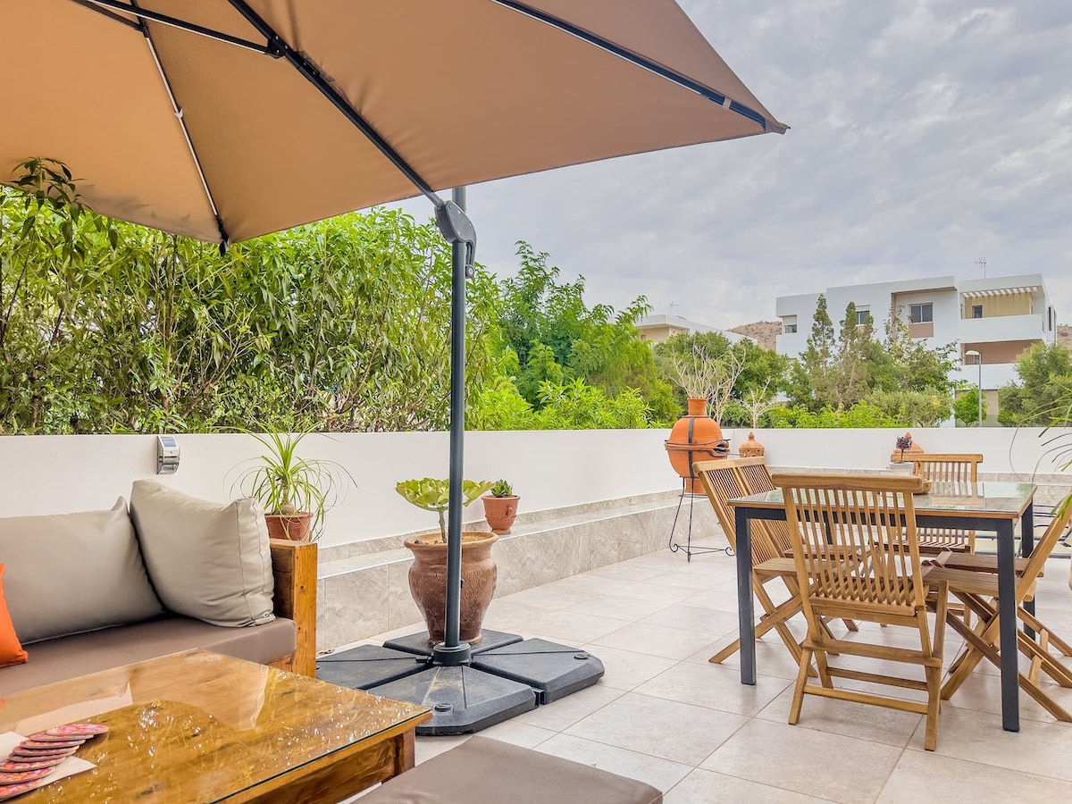 An outdoor terrace is featured, furnished with a shaded lounge area and a dining table. Potted plants add greenery, while a barbecue grill is positioned nearby. The space is tiled and offers a relaxing environment, surrounded by low walls and lush vegetation.