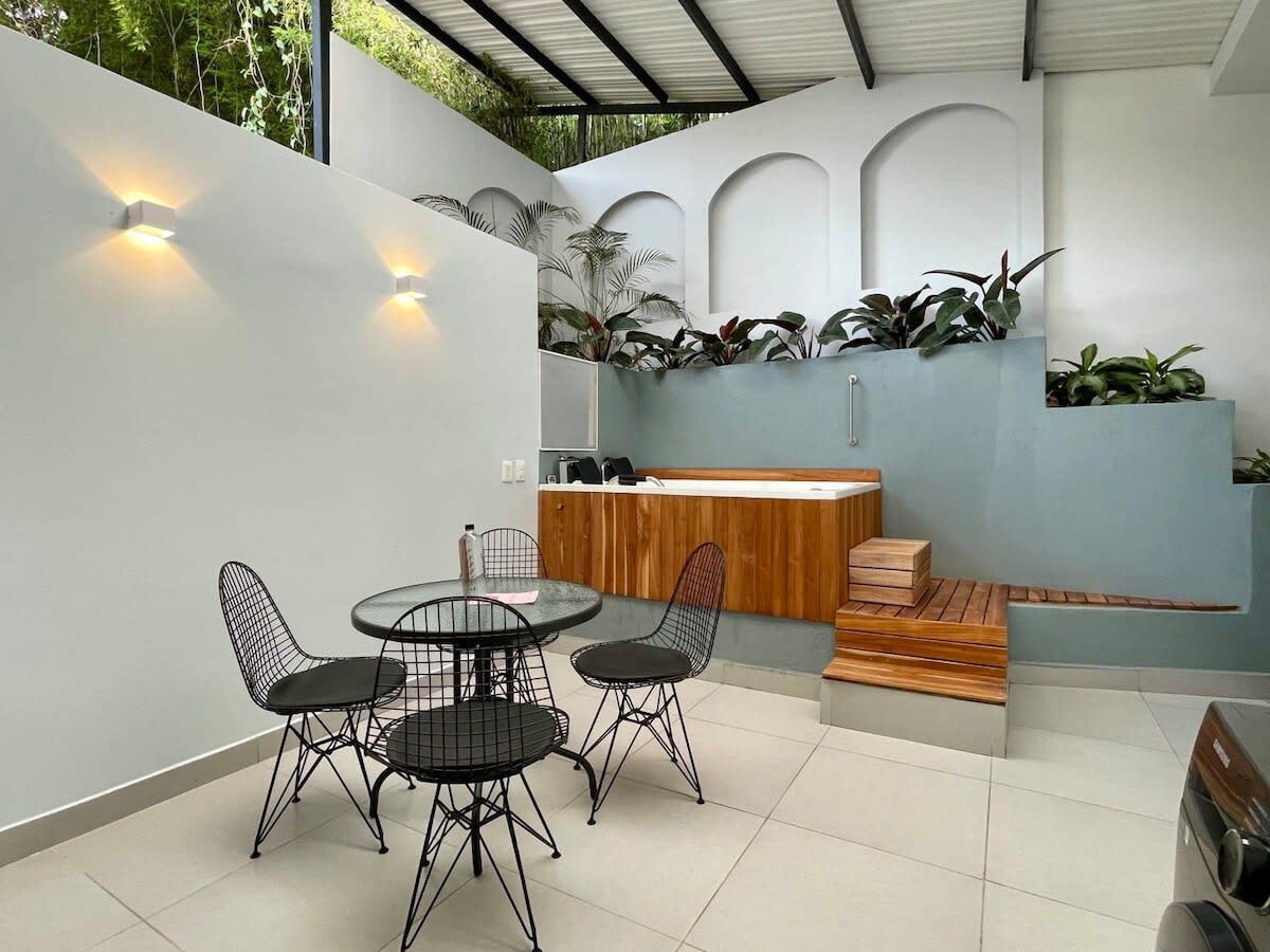 A modern outdoor space features a small dining area with a round glass table and four black wire chairs. In the background, a private jacuzzi is surrounded by lush greenery and decorative plants, providing a peaceful retreat.