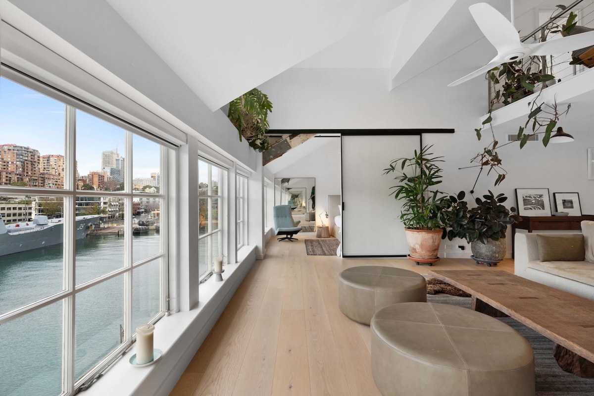 A spacious living area features large windows displaying views of the Harbour and surrounding landscape. Natural light streams in, highlighting potted plants and modern decor. Wooden flooring complements a wooden table and comfortable seating, creating an inviting atmosphere.