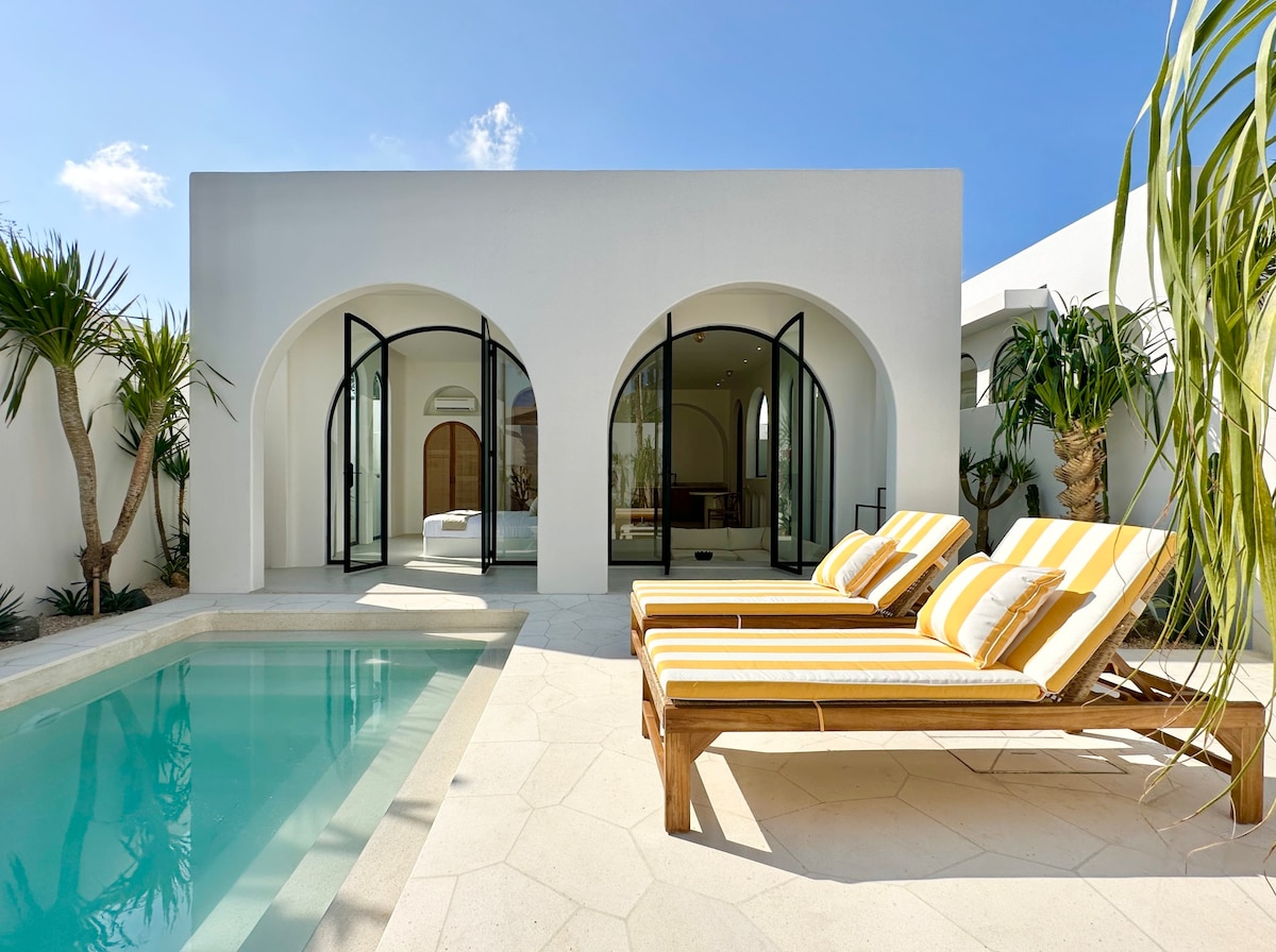 Two sun loungers with yellow and white stripes are positioned beside a private swimming pool. The area is surrounded by tropical plants and features an arched entryway leading to the villa's interior. Clear blue skies and sunlight enhance the inviting outdoor space.